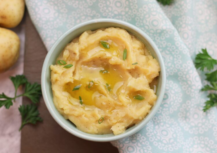 A bowl of mashed potatoes garnished with green onions and a drizzle of olive oil with some potatoes and parsley in the background