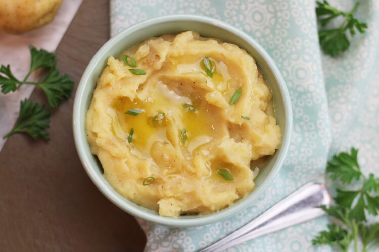 A bowl of mashed potatoes garnished with green onions and a drizzle of olive oil with some potatoes and parsley in the background